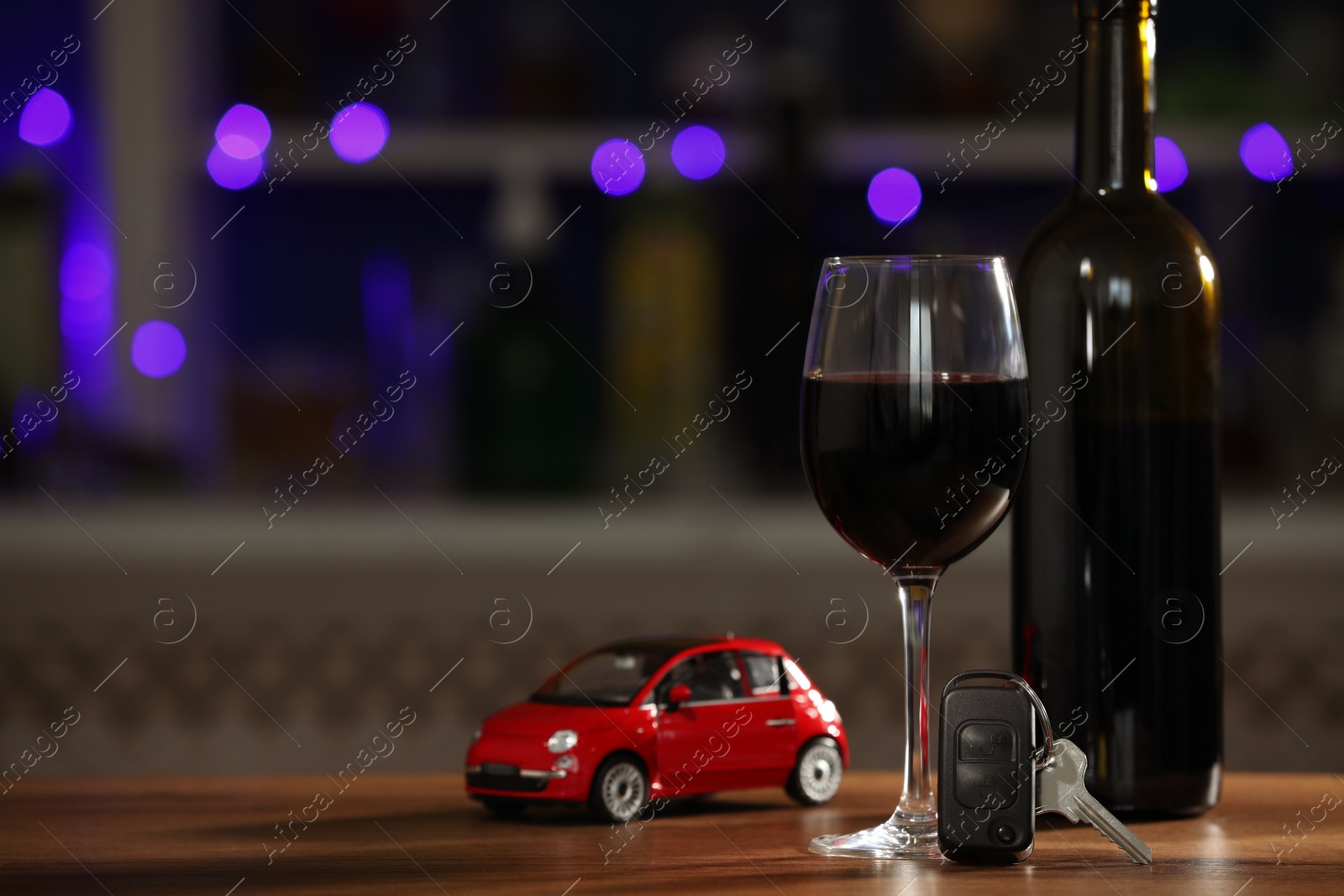 Photo of Car key, glass of alcohol near toy auto on wooden table against blurred lights, space for text. Dangerous drinking and driving
