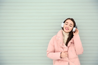 Beautiful young woman listening to music with headphones against light wall. Space for text