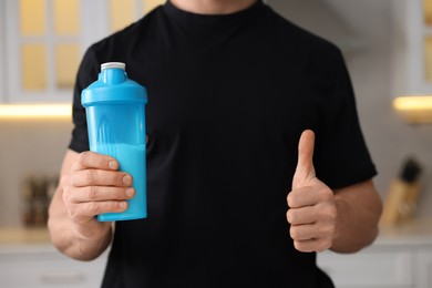 Photo of Young man with shaker of protein showing thumb up in kitchen, closeup