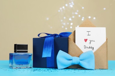 Photo of Happy Father's Day. Card with phrase I Love You, Daddy in envelope, bow tie, perfume and gift box on light blue table, closeup