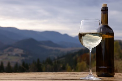 Glass and bottle of tasty wine on wooden table against mountain landscape. Space for text
