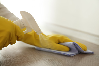 Photo of Person in gloves cleaning wooden table with spray detergent and rag indoors, closeup