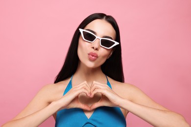 Photo of Beautiful young woman in stylish sunglasses blowing kiss and making heart with hands on pink background