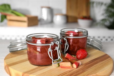 Jars of tasty rhubarb jam, cut stems and strawberry on white table