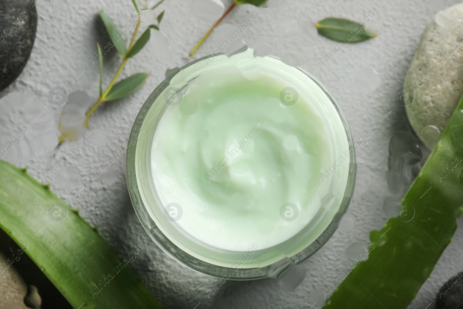 Photo of Flat lay of body cream in jar with aloe and eucalyptus on light grey stone table, view through wet glass