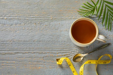 Photo of Cup of herbal diet tea, measuring tape and green leaf on grey wooden table, flat lay with space for text. Weight loss concept