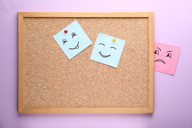 Photo of Cork board and paper notes with drawn faces on violet background. Concept of jealousy