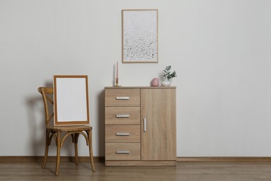 Photo of Wooden chair, chest of drawers, decorative elements and frames in room with light wall, space for text. Interior design
