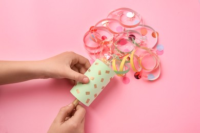 Photo of Woman holding party popper with serpentine and confetti on pink background, top view