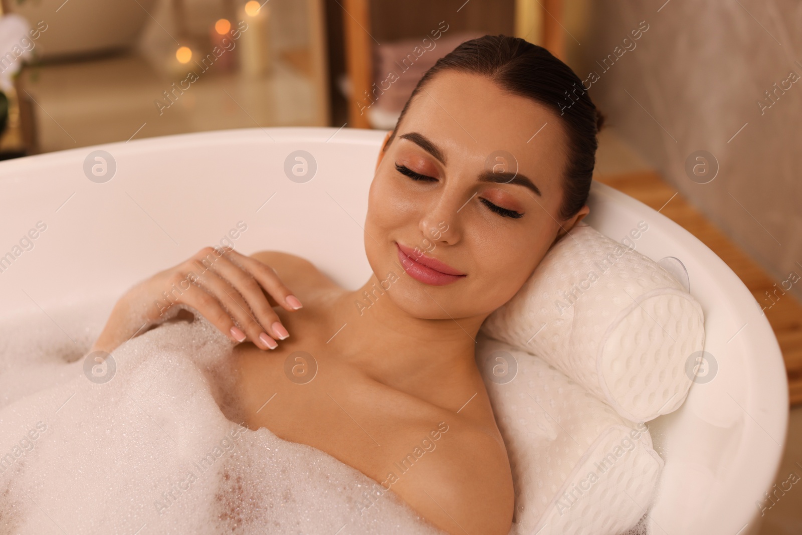 Photo of Young woman using pillow while enjoying bubble bath indoors