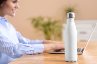 Photo of Young woman with laptop at table indoors, focus on thermo bottle