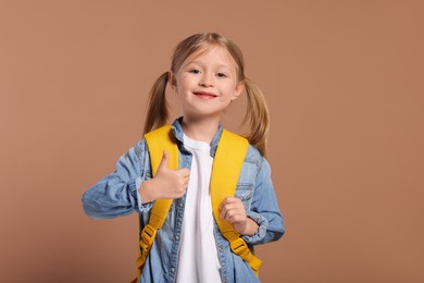 Photo of Happy schoolgirl with backpack showing thumb up gesture on brown background