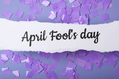 Paper note with phrase APRIL FOOL'S DAY and confetti on lilac background, flat lay