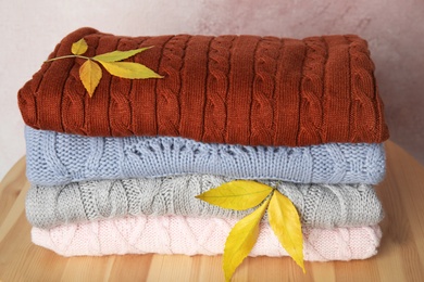 Photo of Stack of warm knitted clothes and autumn leaves on wooden table against light pink background, closeup