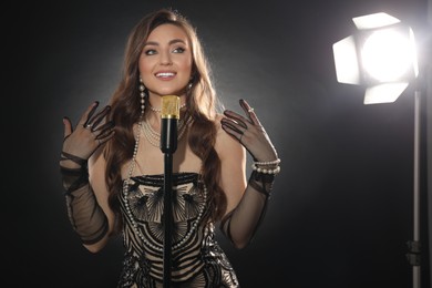 Beautiful young woman in stylish dress with microphone singing on dark background