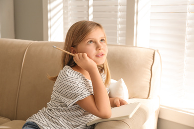 Photo of Thoughtful little girl with pencil and notebook on couch at home