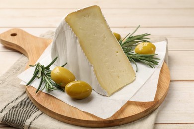 Board with piece of tasty camembert cheese, olives and rosemary on light wooden table, closeup