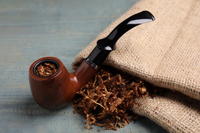 Photo of Smoking pipe, dry tobacco and sackcloth on blue wooden table, closeup