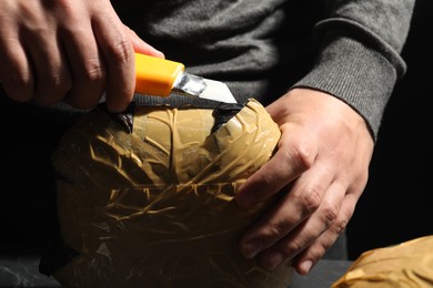 Photo of Smuggling and drug trafficking. Man opening package of narcotics with box cutter on black background, closeup