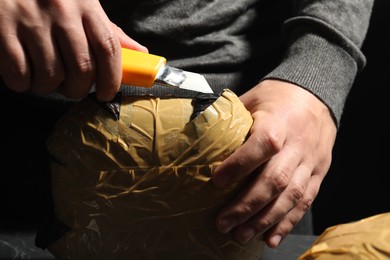 Smuggling and drug trafficking. Man opening package of narcotics with box cutter on black background, closeup