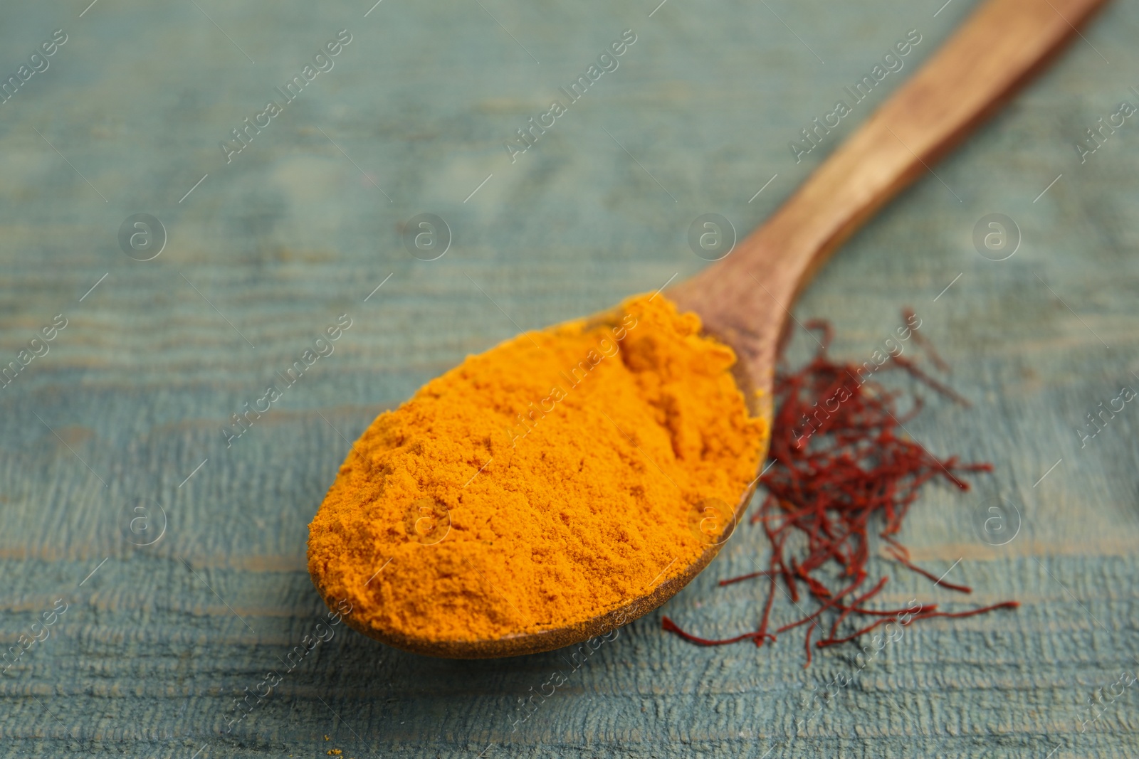 Photo of Spoon with saffron powder and dried flower stigmas on light blue wooden table, closeup