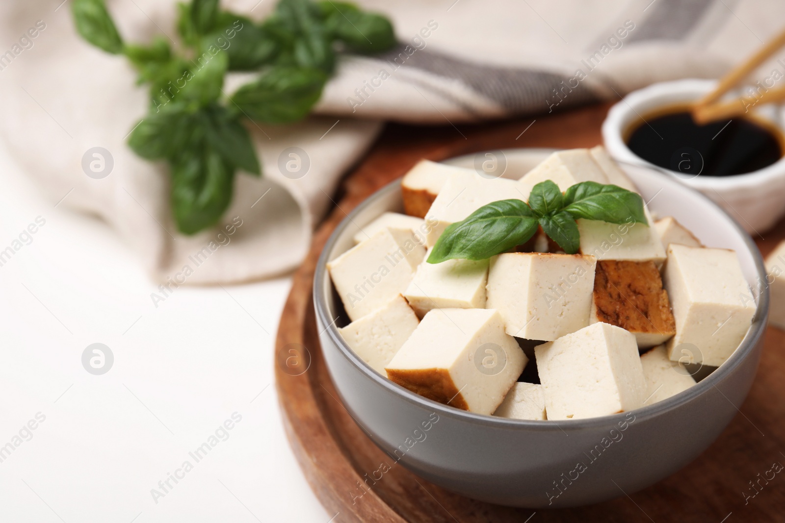 Photo of Wooden tray with bowl of smoked tofu cubes, basil and soy sauce on white table, closeup. Space for text