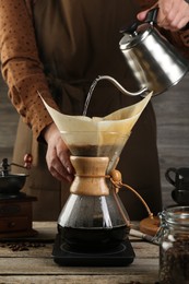 Woman pouring hot water into glass chemex coffeemaker with paper filter at wooden table, closeup
