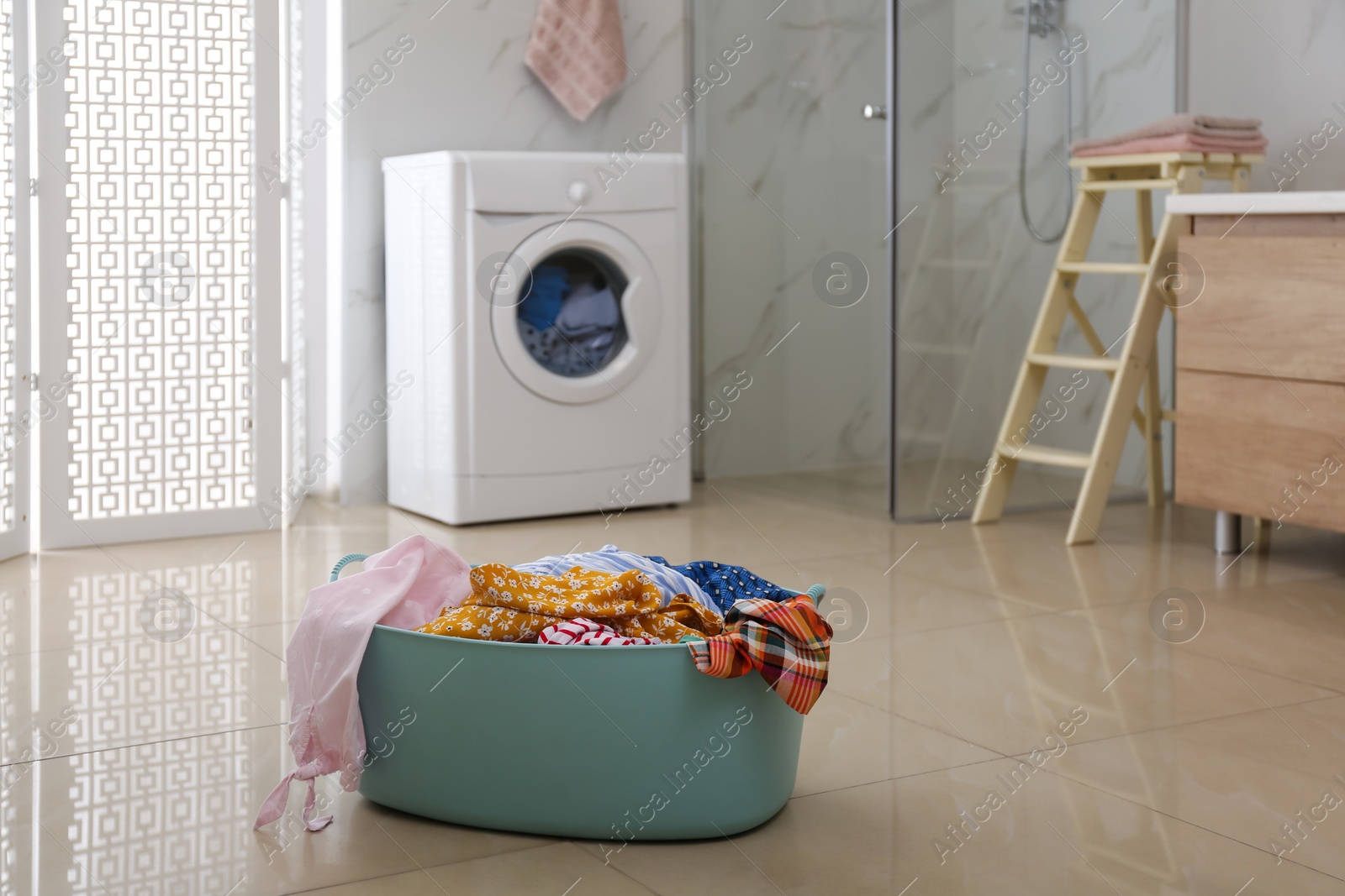 Photo of Plastic laundry basket full of different clothes on floor in bathroom