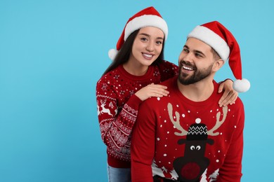 Happy young couple in Christmas sweaters and Santa hats on light blue background. Space for text