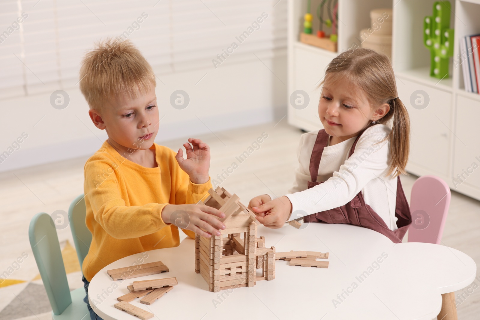 Photo of Little boy and girl playing with wooden house at white table indoors. Children's toys