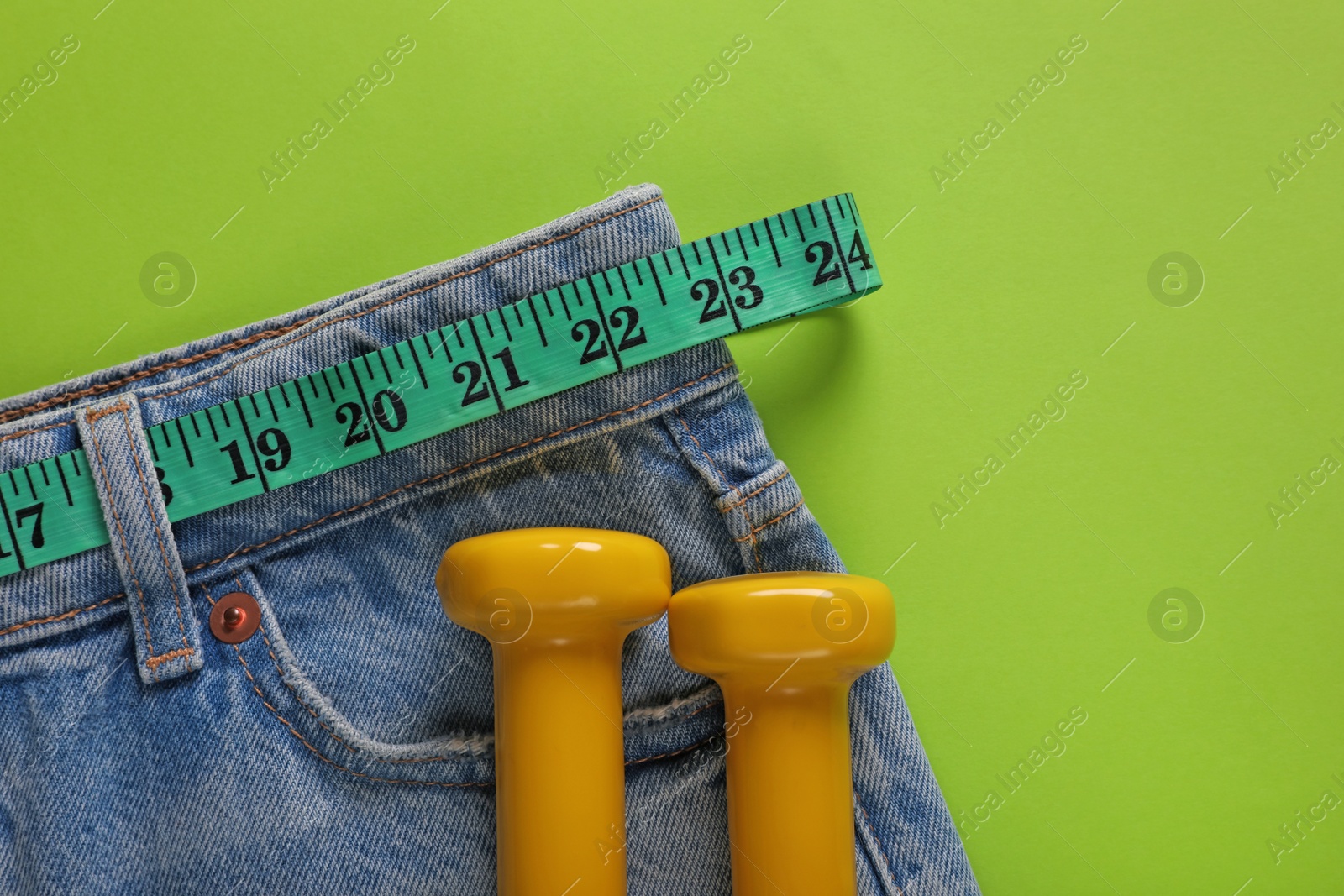 Photo of Jeans, dumbbells and measuring tape on light green background, flat lay with space for text. Weight loss concept