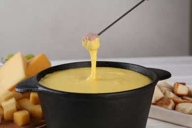 Dipping piece of ham into fondue pot with tasty melted cheese at table against gray background, closeup