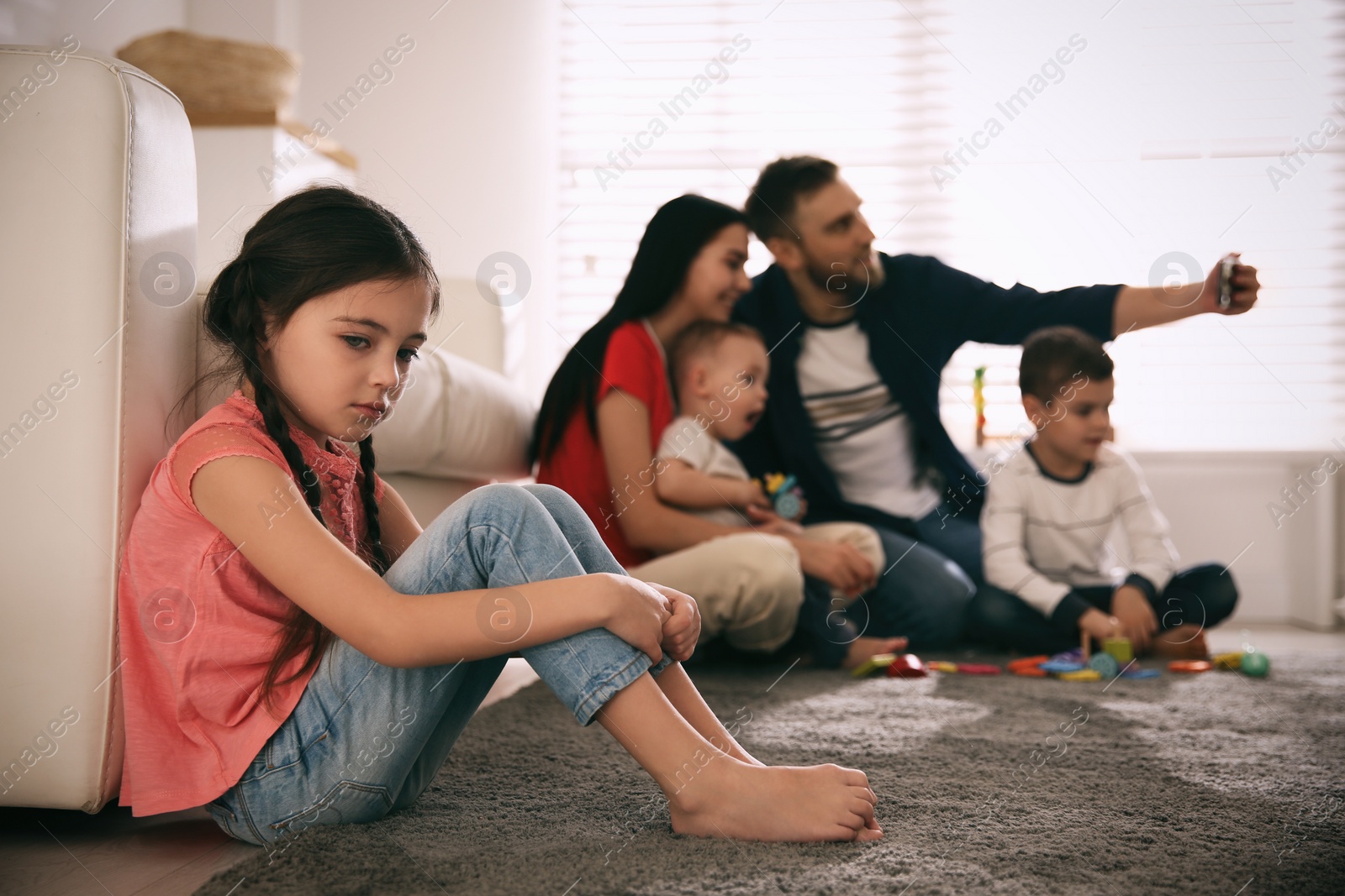 Photo of Unhappy little girl feeling jealous while parents taking selfie with other children at home