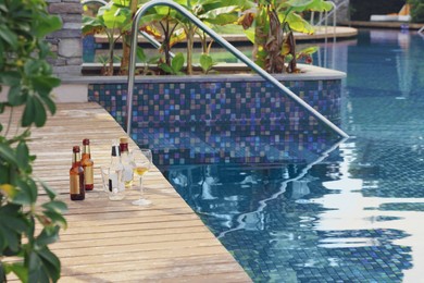 Photo of Wooden deck with bottles of drink and glasses near outdoor swimming pool. Luxury resort