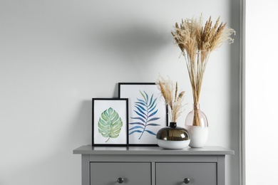 Reed's blossom in glass vases and pictures on grey cabinet indoors. Space for text