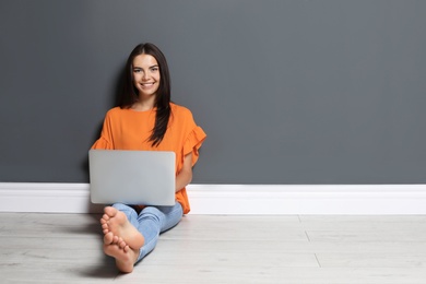 Photo of Young woman with modern laptop sitting on floor near grey wall