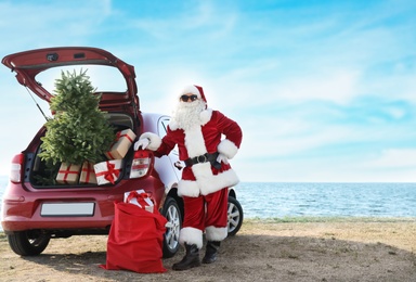 Authentic Santa Claus near red car with gift boxes and Christmas tree on beach