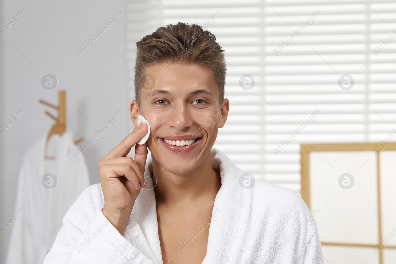 Photo of Handsome young man cleaning face with cotton pad in bathroom