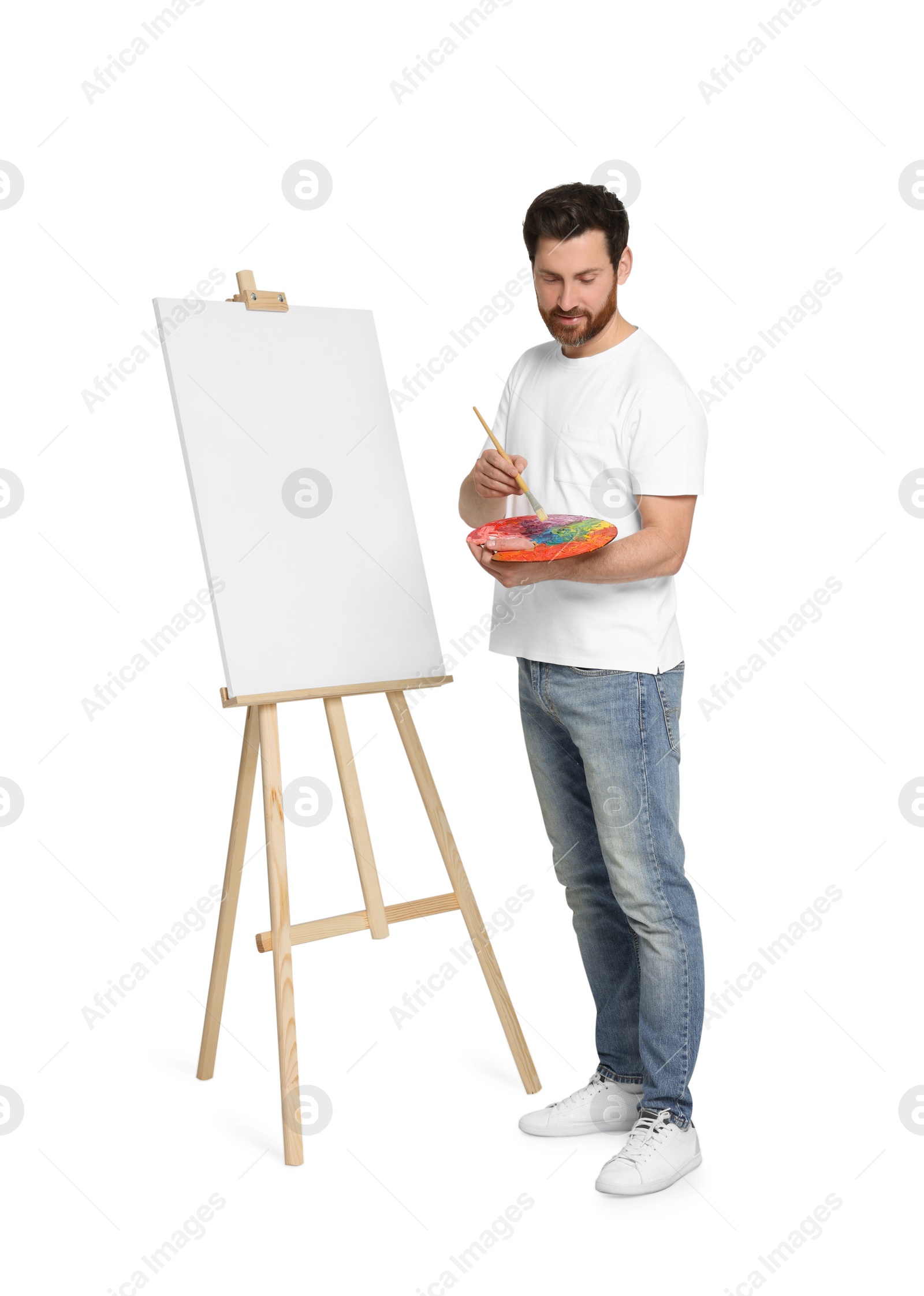 Photo of Man with brush and artist`s palette painting against white background. Using easel to hold canvas