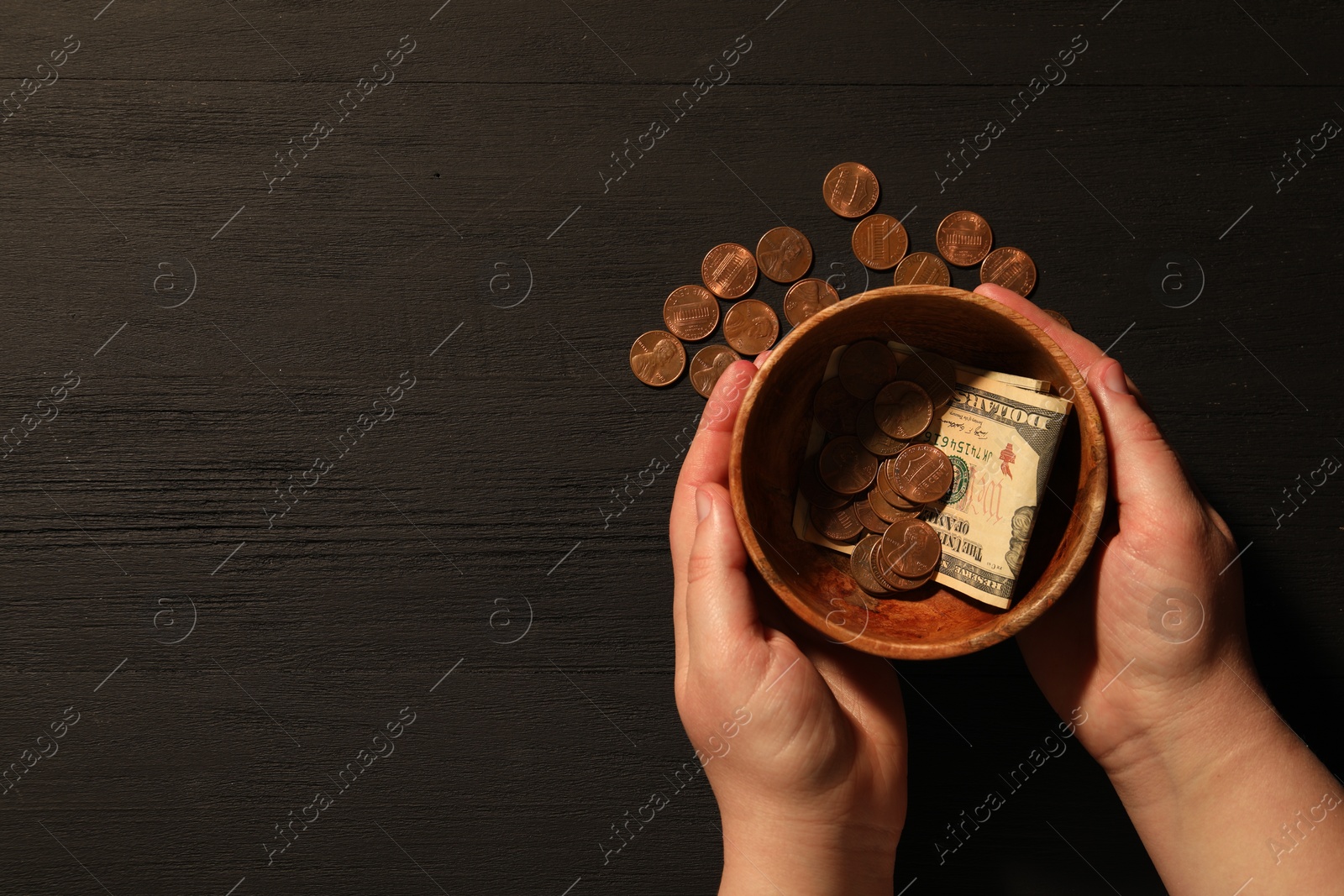 Photo of Donate and give concept. Woman holding bowl with coins and dollar banknotes at black wooden table, top view. Space for text
