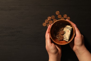 Donate and give concept. Woman holding bowl with coins and dollar banknotes at black wooden table, top view. Space for text