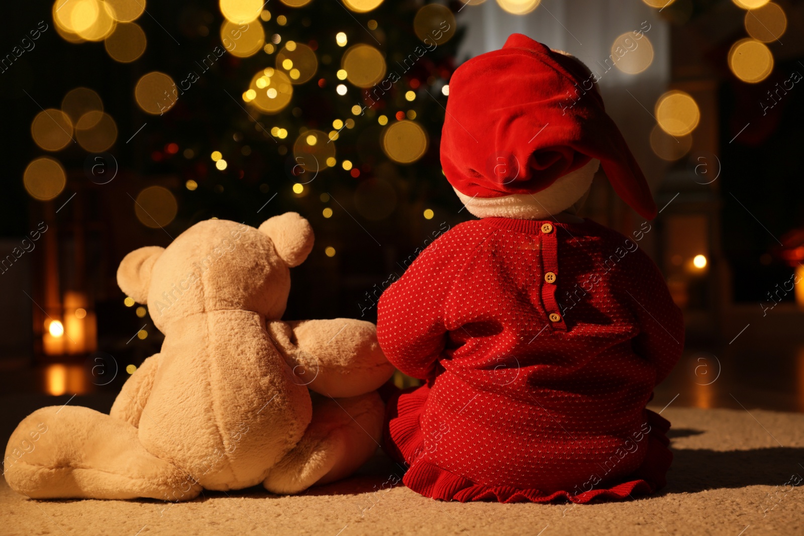 Photo of Baby wearing Santa hat with teddy bear in room decorated for Christmas, back view