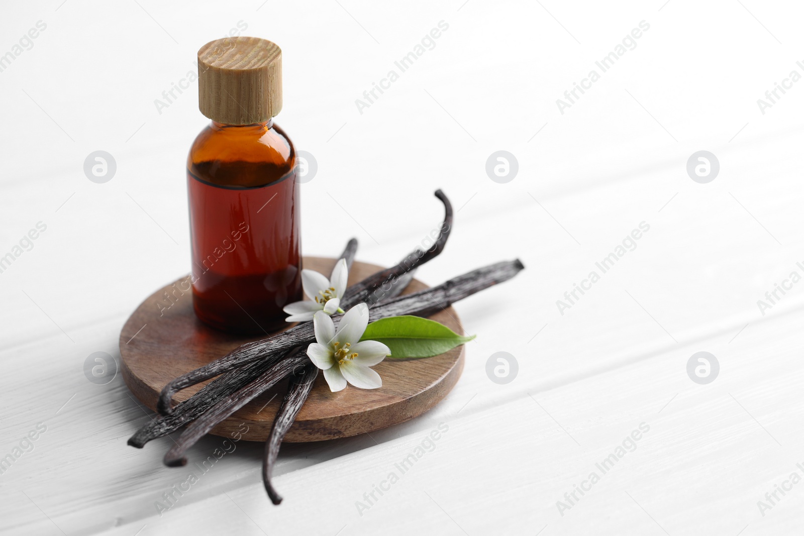 Photo of Vanilla pods, flowers, leaf and bottle with essential oil on white wooden table, closeup. Space for text