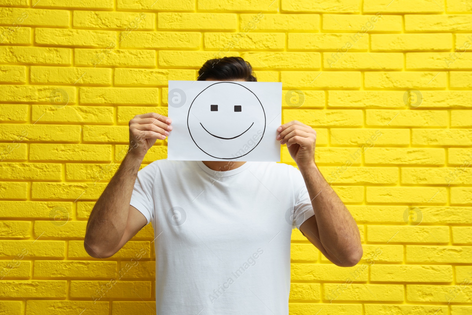 Photo of Man hiding emotions using card with drawn smiling face near yellow brick wall