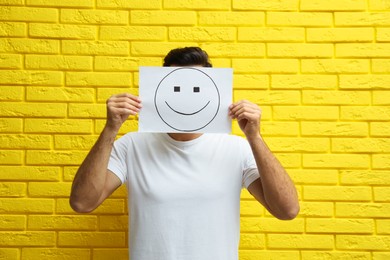 Photo of Man hiding emotions using card with drawn smiling face near yellow brick wall