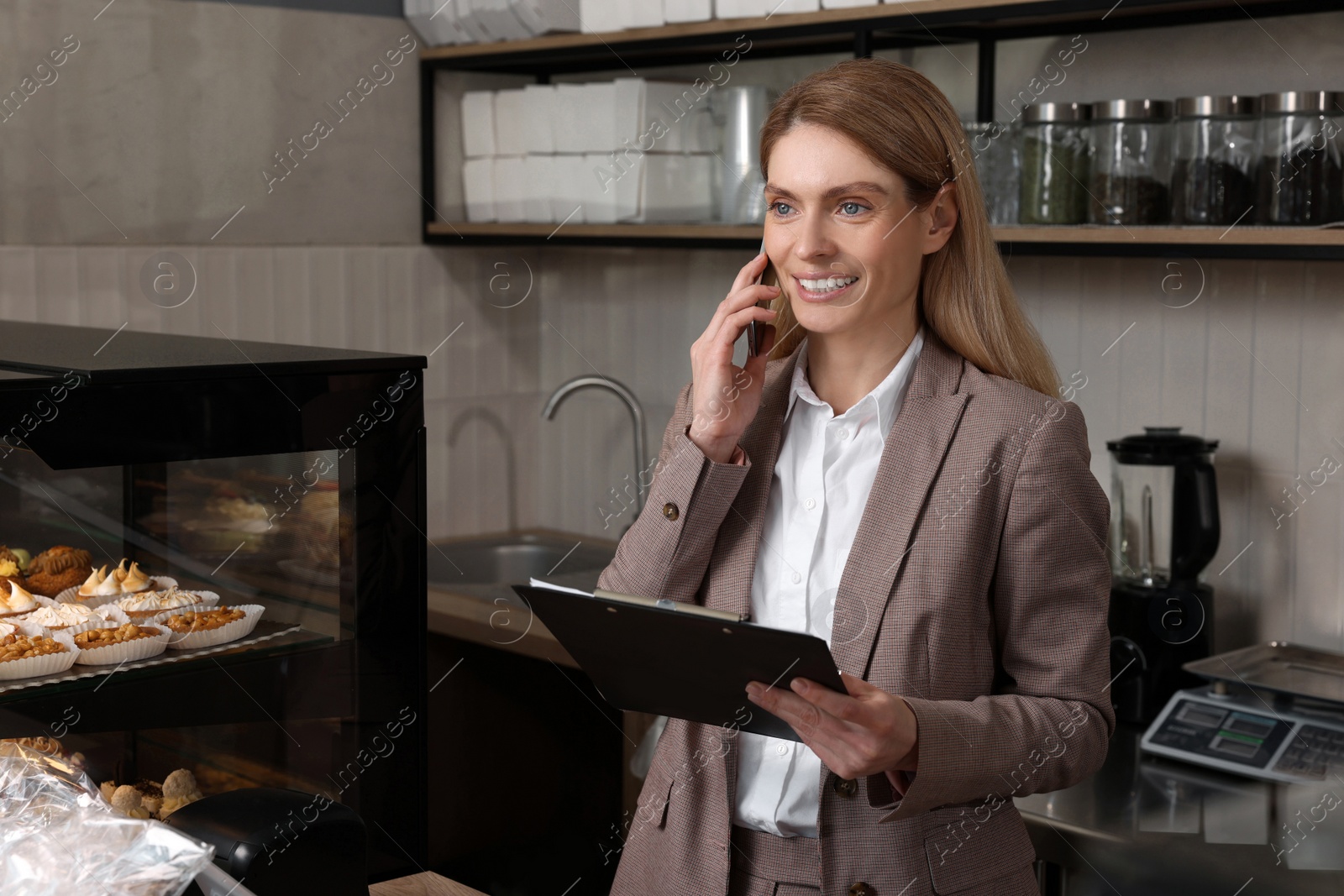 Photo of Happy business owner with clipboard talking on phone in bakery shop