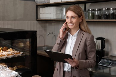 Photo of Happy business owner with clipboard talking on phone in bakery shop