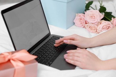 Woman using laptop near gift and rose flowers on bed in room, closeup. Happy Birthday