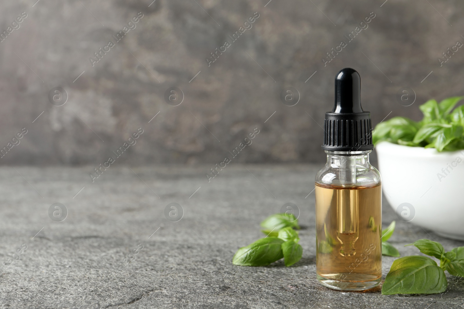 Photo of Glass bottle of basil essential oil and leaves on grey stone table. Space for text