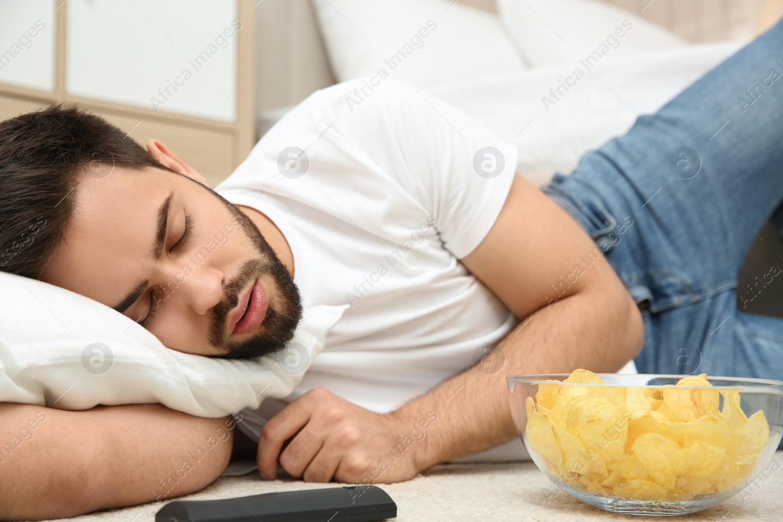 Photo of Lazy young man with bowl of chips and TV remote sleeping on floor at home